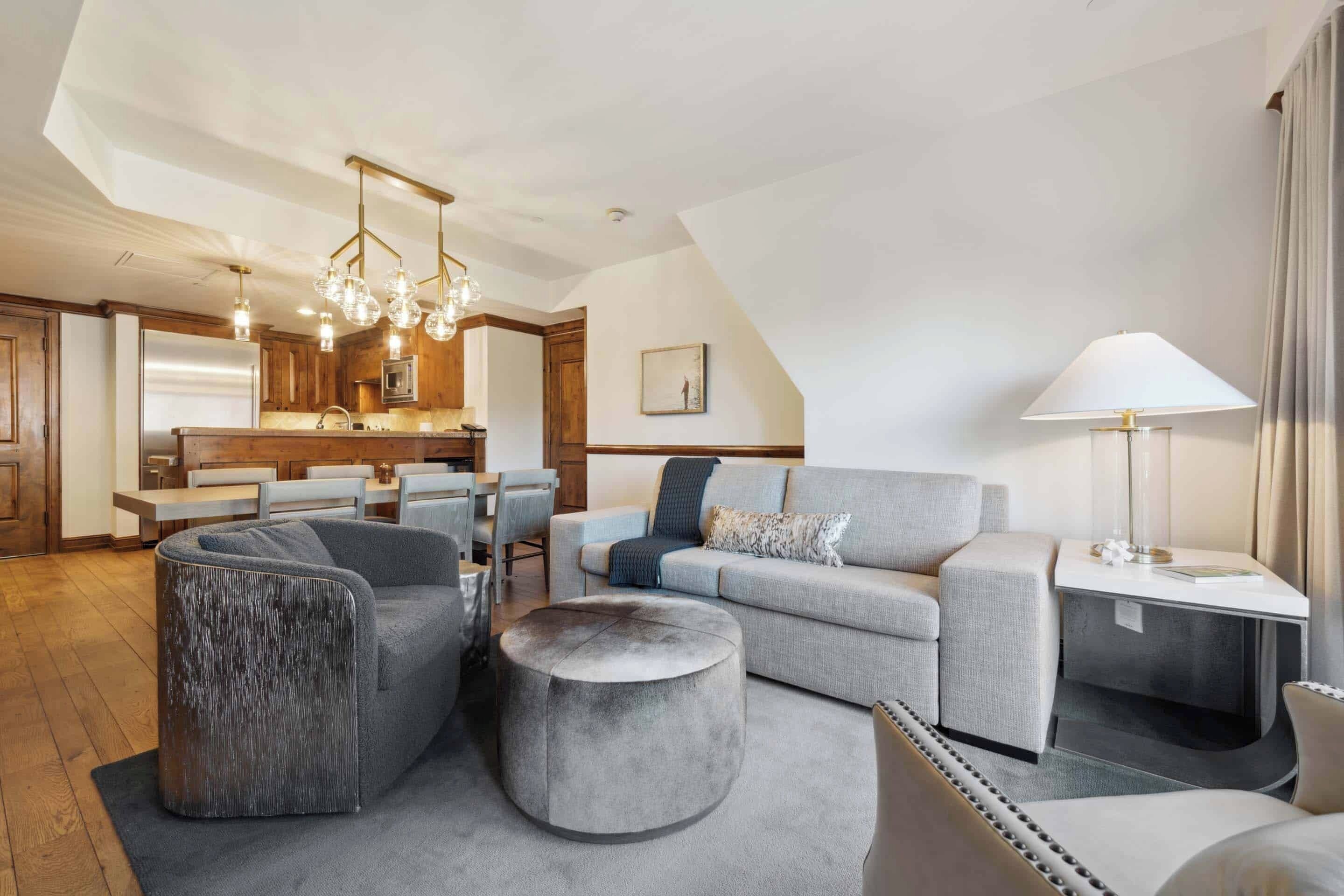 Een moderne woonkamer met een grijze bank, een poef en een fauteuil, een eethoek met een houten tafel en stoelen, en een keuken met een koelkast en hanglampen.