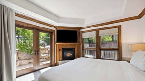 Un dormitorio con una cama grande, una chimenea, un televisor y un balcón con un muro de piedra y vegetación afuera.