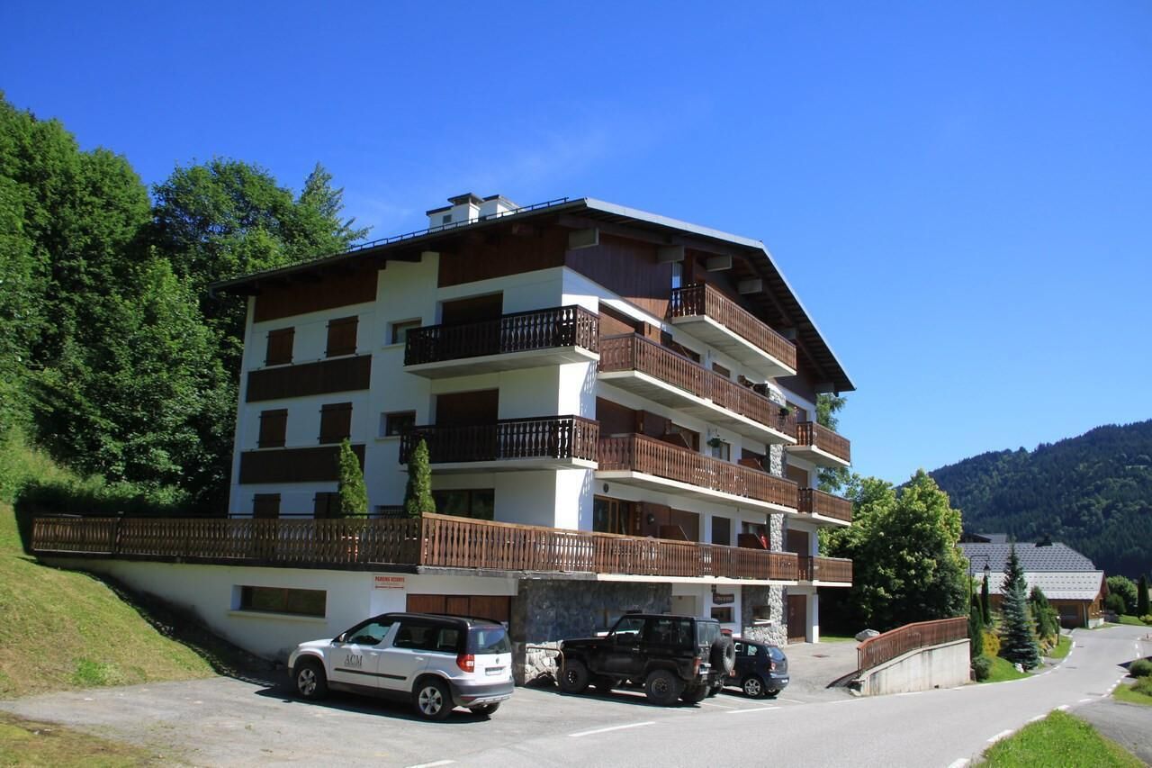 A multi-story building with balconies, a parking area, and a mountainous backdrop.