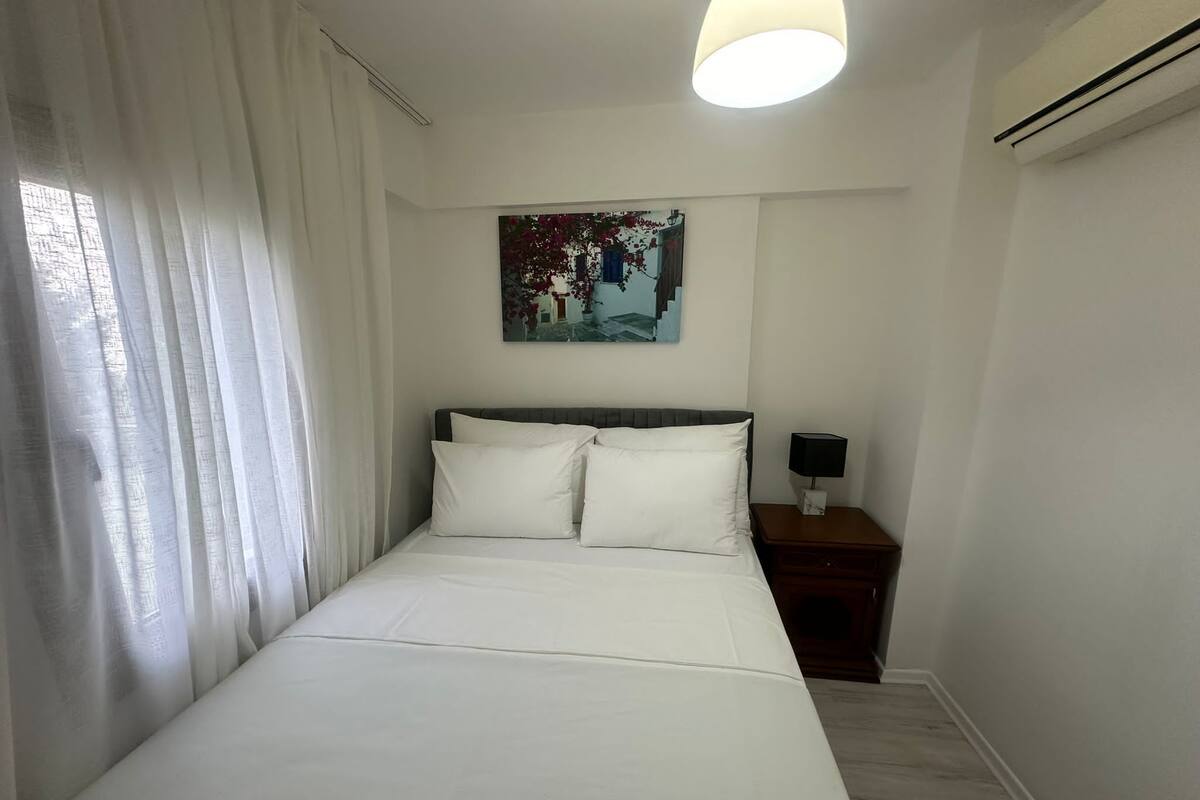 A neatly made bed with white linens in a room with a single nightstand, a painting on the wall, and a window with sheer curtains.