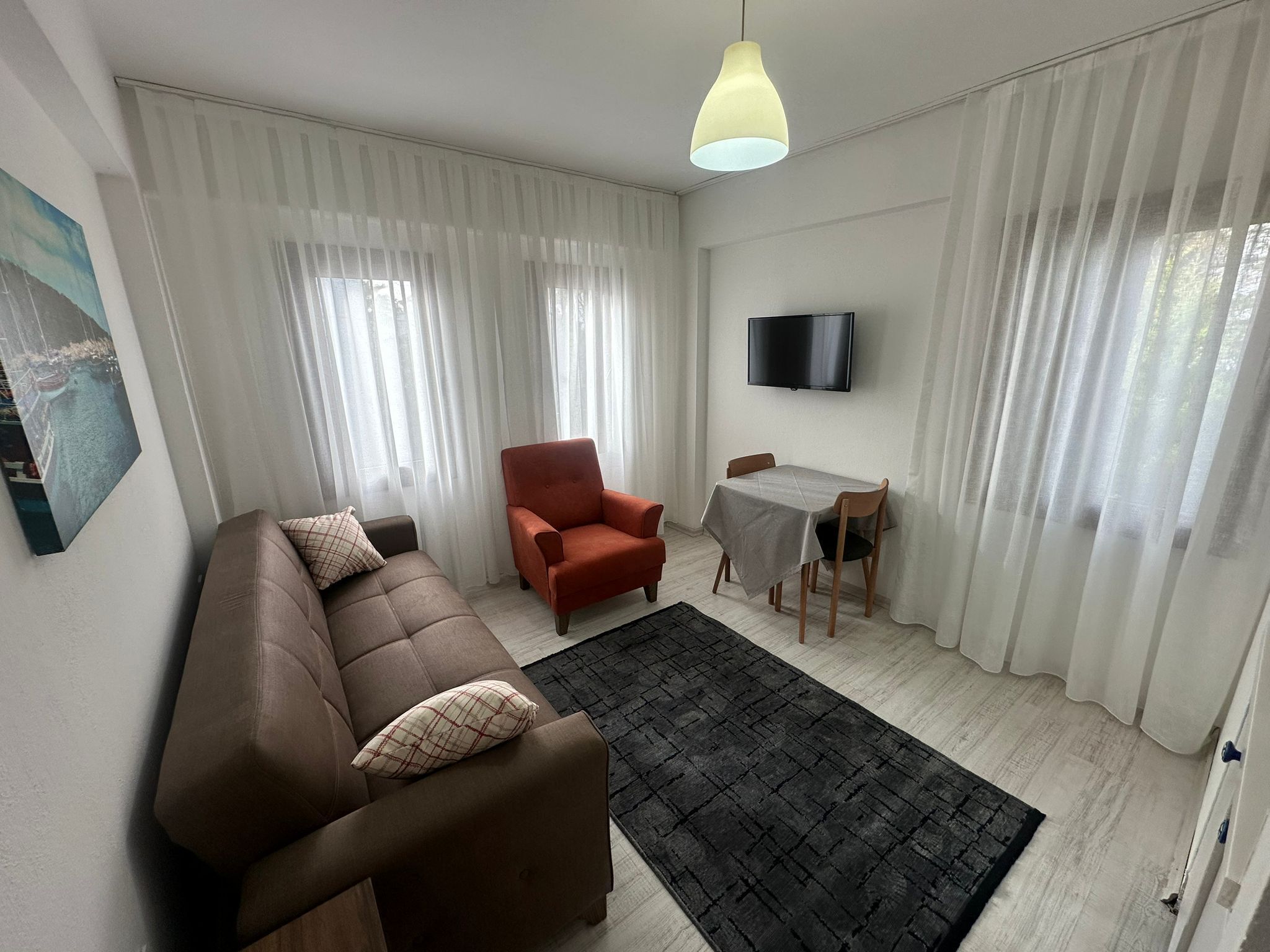 A living room with a brown sofa, a red armchair, a dining table with two chairs, a black rug, and a TV on the wall.