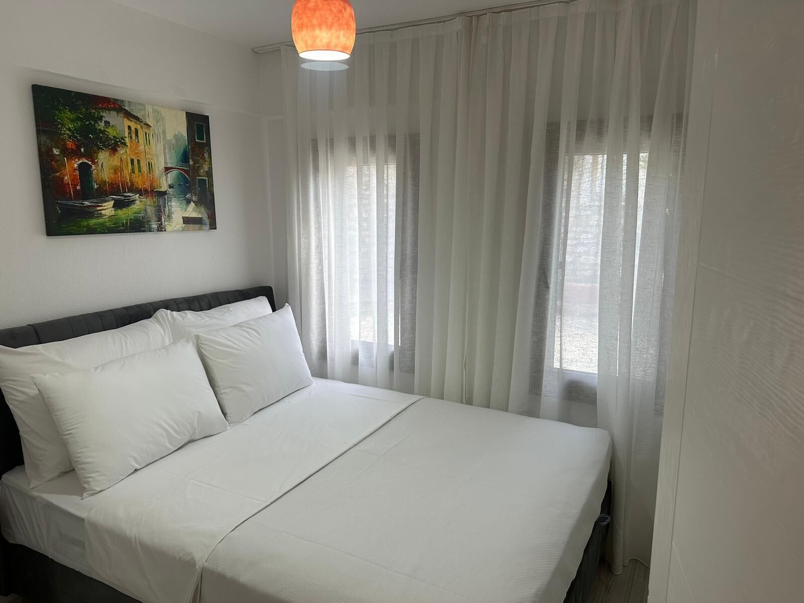 Ein ordentlich bezogenes Bett mit weißen Leinen in einem Zimmer mit einem großen Fenster, das mit transparenten Vorhängen verziert ist, einem Gemälde an der Wand und einer Pendelleuchte.