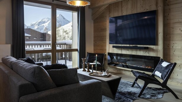 Un soggiorno moderno con una grande TV, un camino e vista sulle montagne innevate.