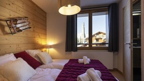 Hotelkamer met houten hoofdeinde, bed met wit en rode beddengoed, paarse deken, en raam met uitzicht op een berg en gebouwen.