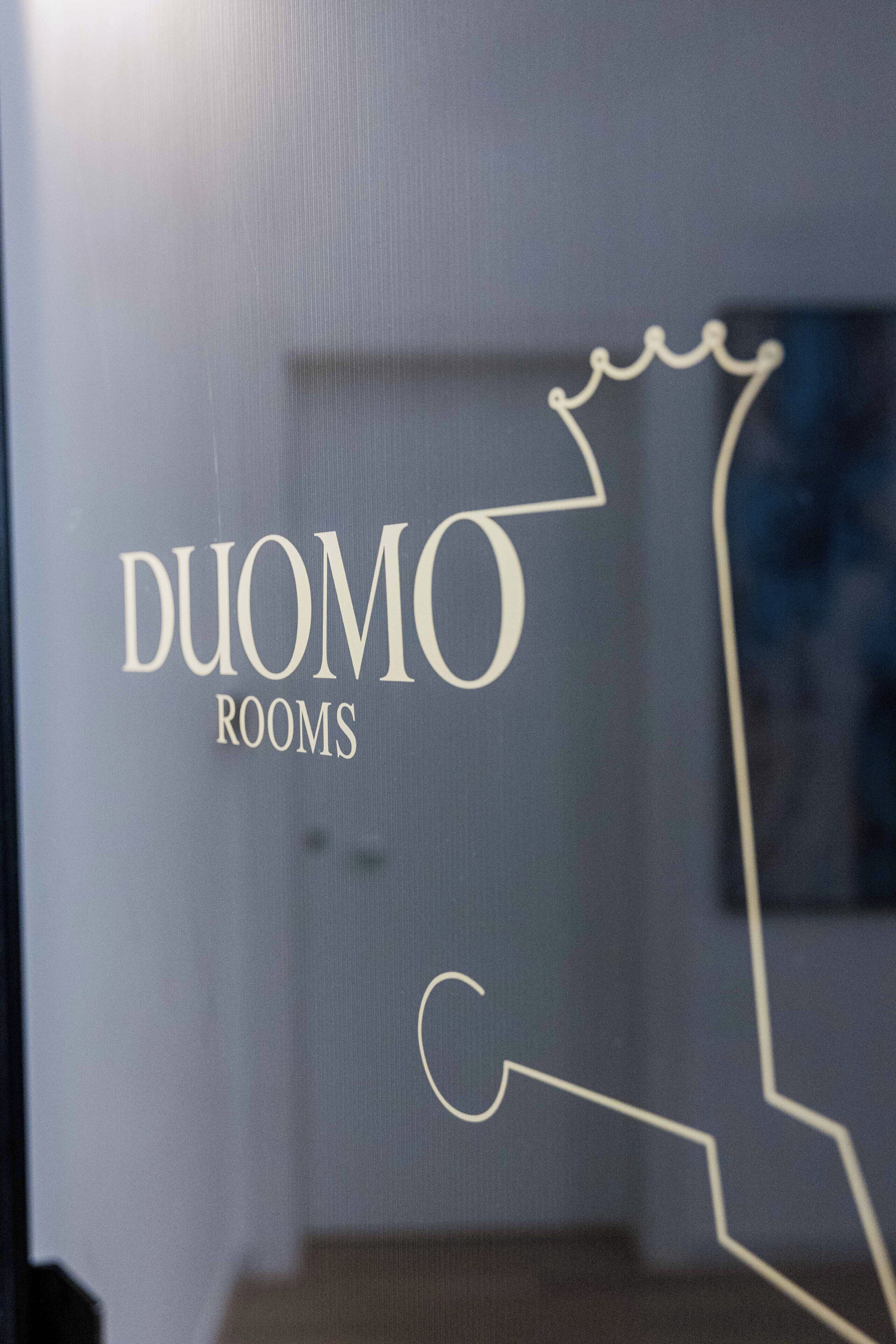 A glass window with the text "DUOMO ROOMS" and a simple line drawing of a building outline.