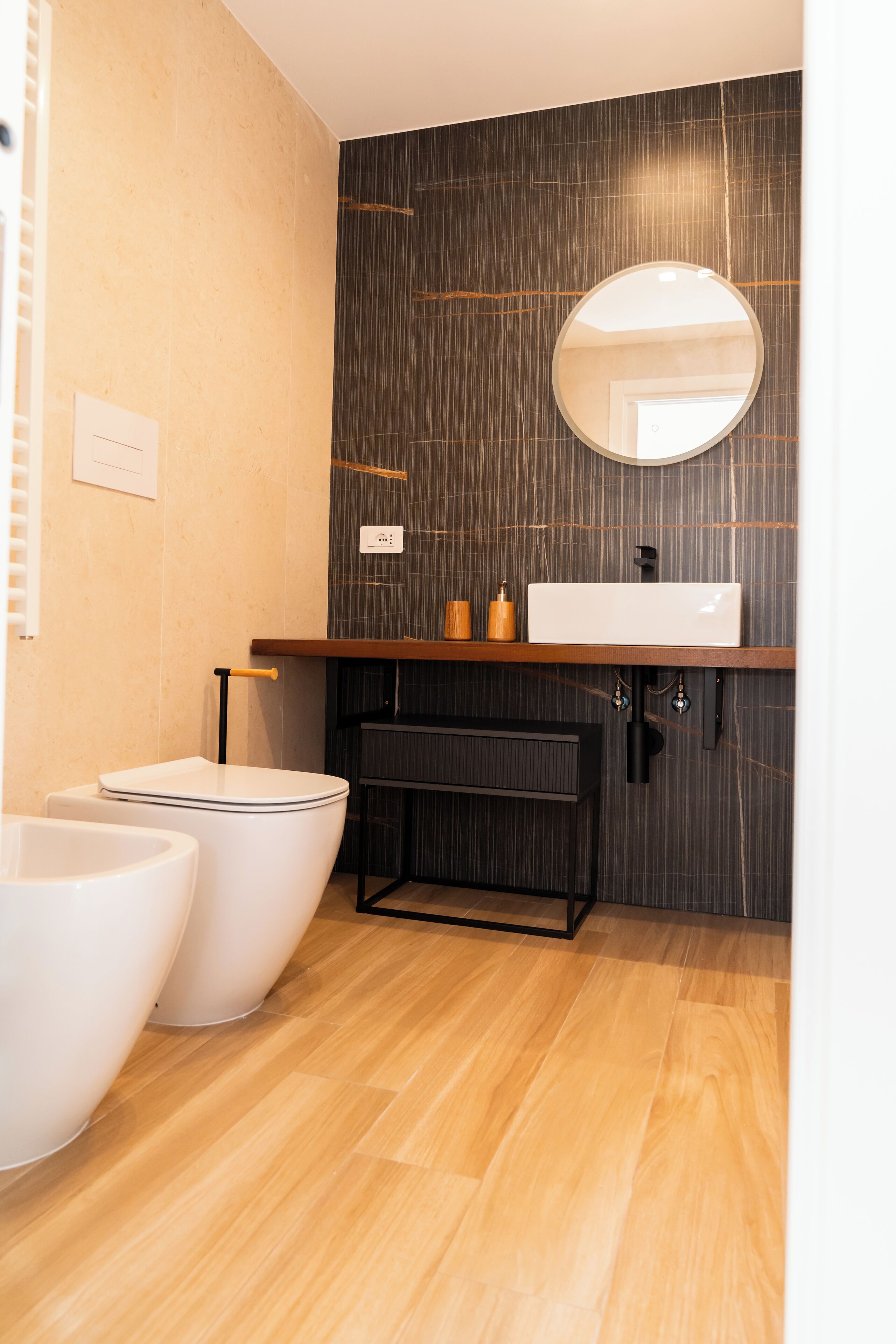 Een moderne badkamer met een houten wastafelmeubel, een ronde spiegel en twee witte toiletten.