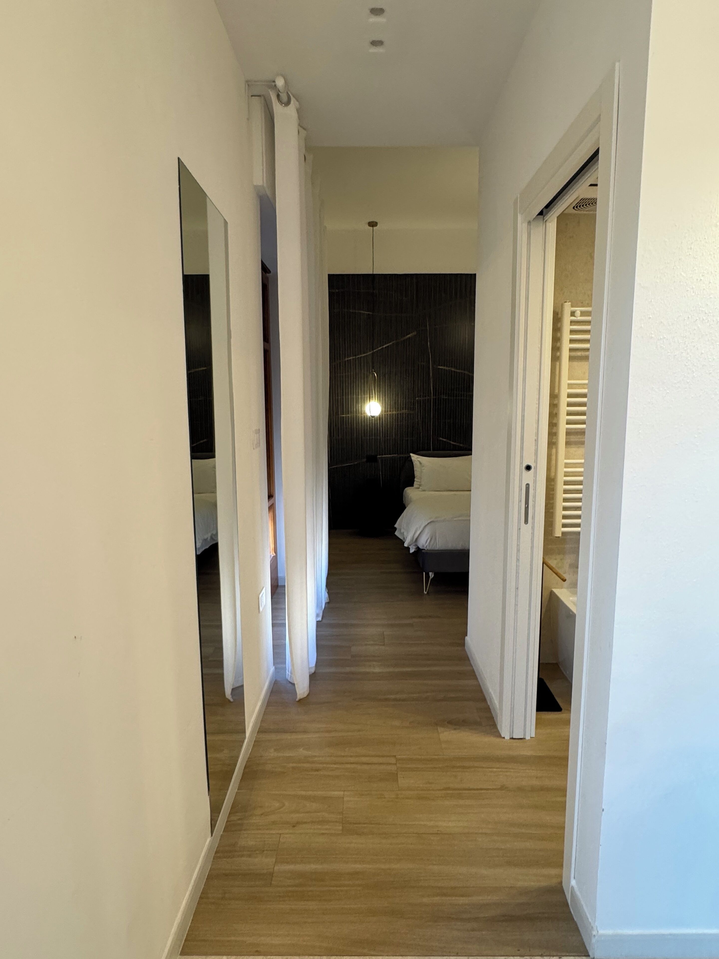 A narrow hallway with wooden flooring leading to a bedroom with a bed and a nightstand.