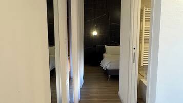 A narrow hallway with wooden flooring leading to a bedroom with a bed and a nightstand.