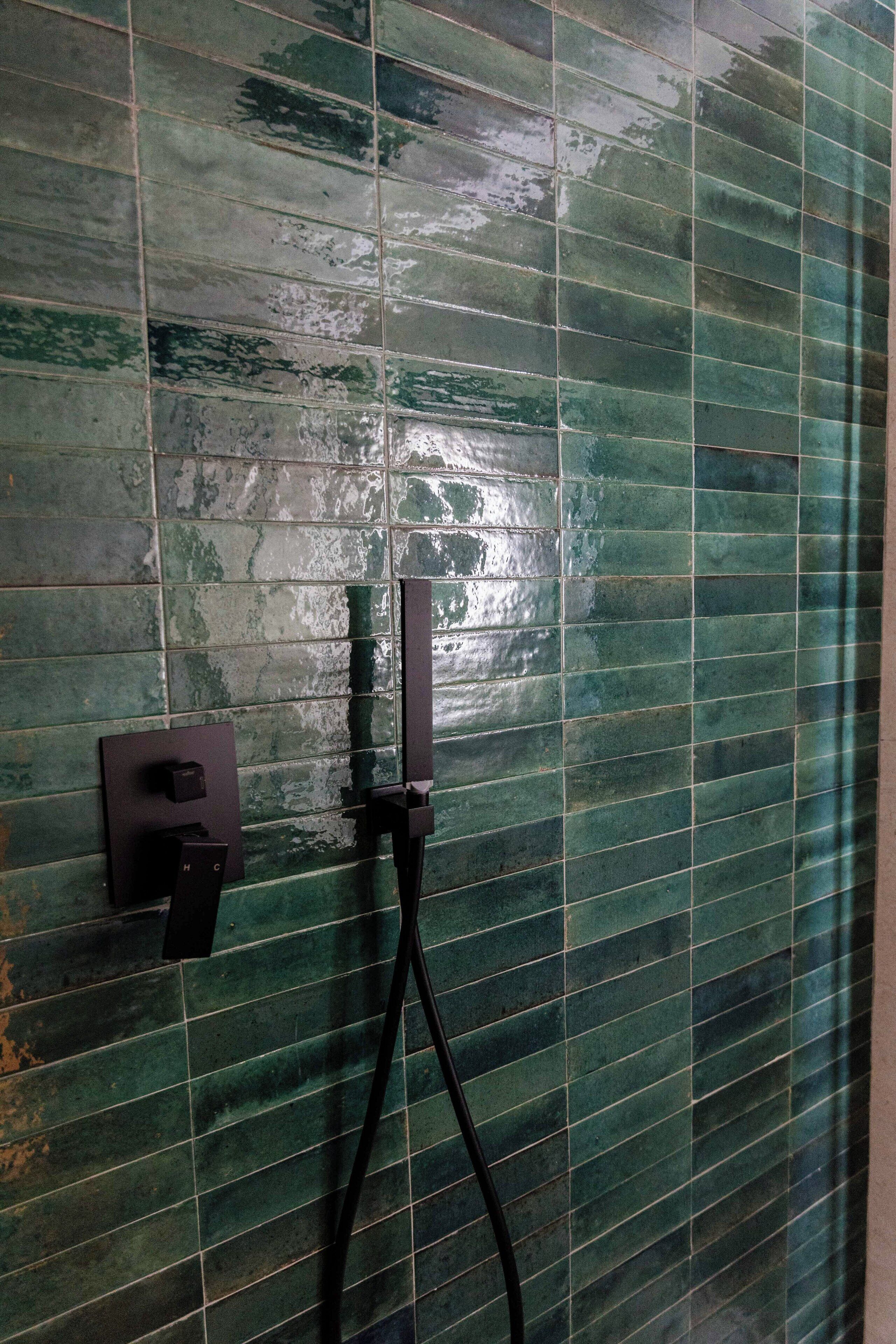 A bathroom with green tiled walls and a shower area with a black hose and control panel.