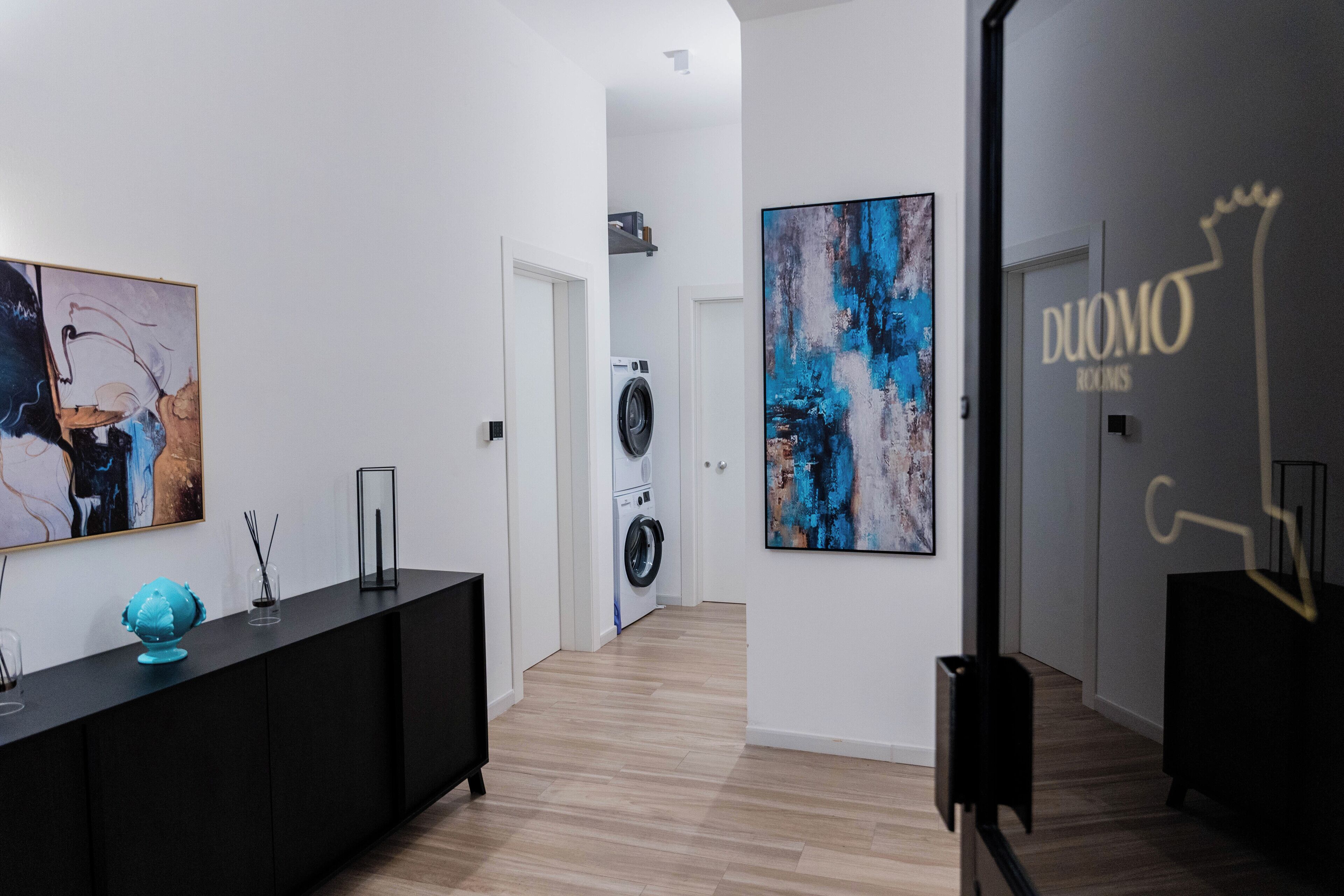 A hallway with a black console table, a large abstract painting, a washing machine, and a sign reading "DUOMO ROOMS".