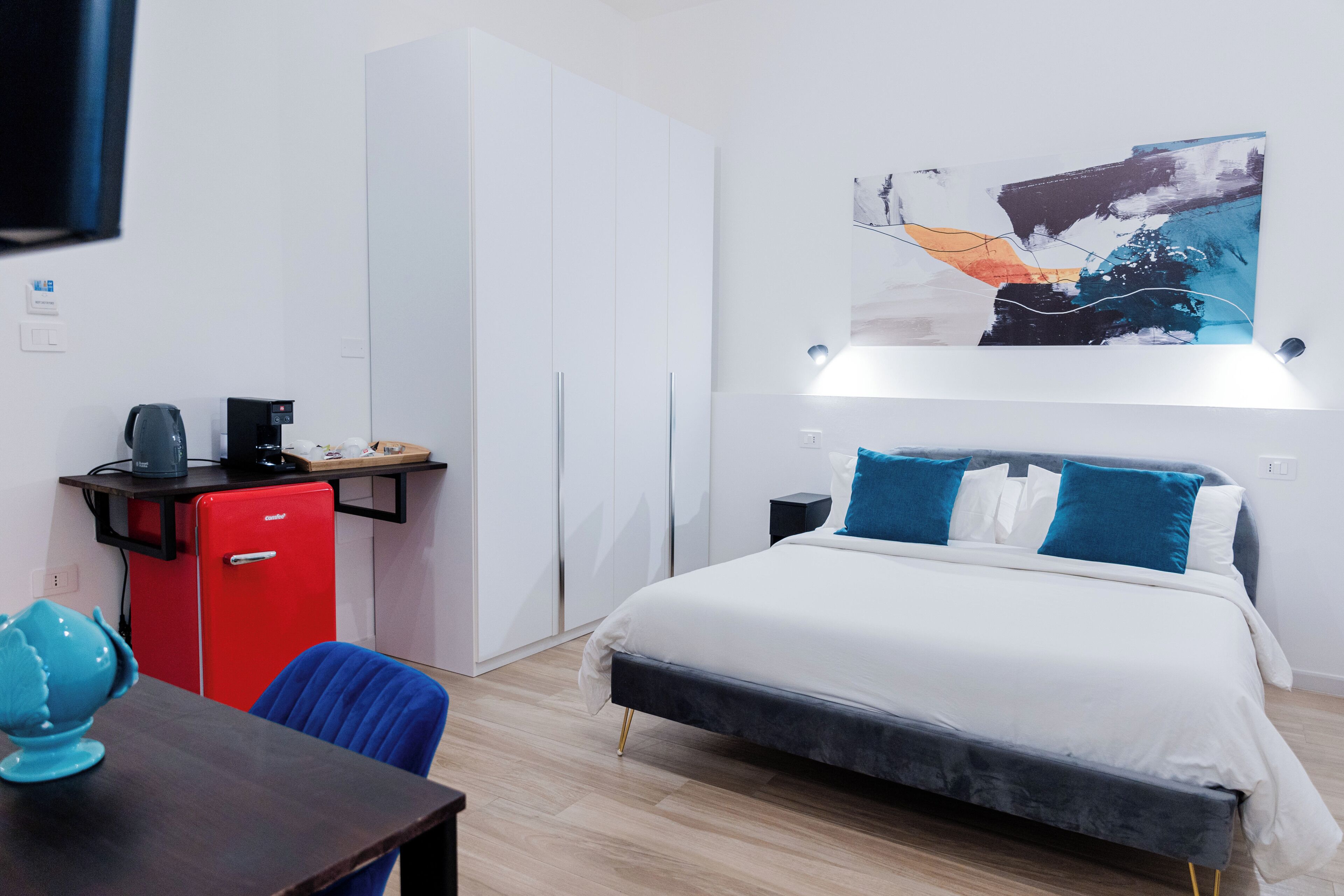 Une chambre d’hôtel moderne équipée d’un lit, d’un bureau avec un mini-réfrigérateur rouge, d’une machine à café et d’une chaise bleue.