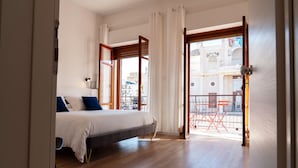 A bedroom with a bed, a view of the street, and a balcony with chairs.