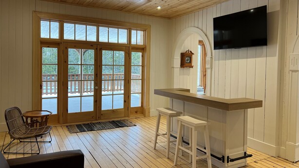 Een ruime kamer met een houten plafond, grote ramen, een bar met krukken en een televisie aan de muur.