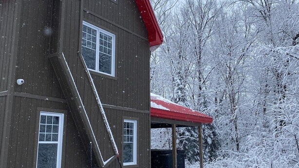 Una casa de madera de dos pisos con techo rojo y un porche cubierto. Se ve nieve en el techo, las ventanas y los árboles circundantes.