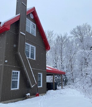 Een houten huis met twee verdiepingen, voorzien van een rood dak en een overdekte veranda. Er ligt sneeuw op het dak, de ramen en de omliggende bomen.