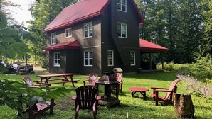 Una casa de dos pisos con techo rojo y un porche. Hay mesas y sillas de picnic en el jardín delantero.