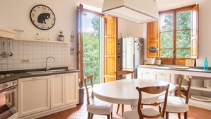 A kitchen with white cabinets, a round dining table, and chairs. There is a clock on the wall and a window with a view of greenery. - Wonderful apartment in Villa with nice garden and terrace. Lovely neighborhood   (Firenze)
