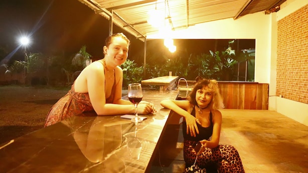 Two people sitting at a table with a glass of wine, in an outdoor setting with a brick wall and plants in the background. - Asha Hostel (La Fortuna)
