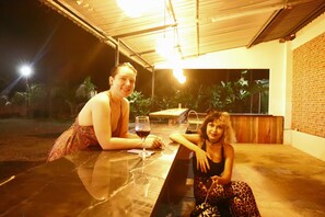 Two people sitting at a table with a glass of wine, in an outdoor setting with a brick wall and plants in the background. - Asha Hostel (La Fortuna)