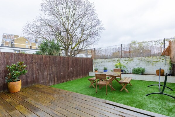 Un jardin avec une terrasse en bois, une pelouse verdoyante, une table en bois entourée de chaises et un mur en briques blanches.