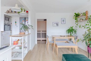 Une cuisine moderne avec des armoires blanches, un plan de travail en bois et un four encastré. On y trouve un coin repas avec une table et des chaises en bois, ainsi qu’un pouf vert.