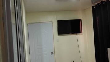Une chambre à coucher avec un lit, une porte, un téléviseur fixé au mur et une fenêtre avec des rideaux.