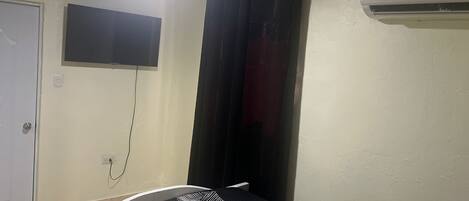 A bedroom with a bed, a TV mounted on the wall, a window with curtains, and an air conditioning unit.