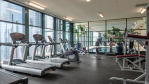 A modern gym with treadmills, elliptical machines, and weight equipment. Large windows offer views of the cityscape. - Elegant 4B 2B Unit in Melbourne CBD w/ Parking! (Melbourne)