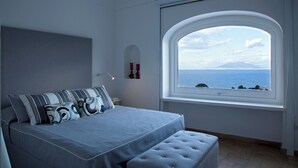 A bedroom with a large bed, a window with a view of the sea, and a small table with a lamp. - Villa Fortuna by MyVillaCollection (Capri)
