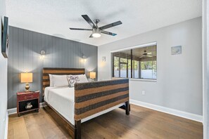 Ein Schlafzimmer mit einem Bett, einem Deckenventilator, einem Fenster mit Jalousien und einem Nachttisch mit Lampe.
