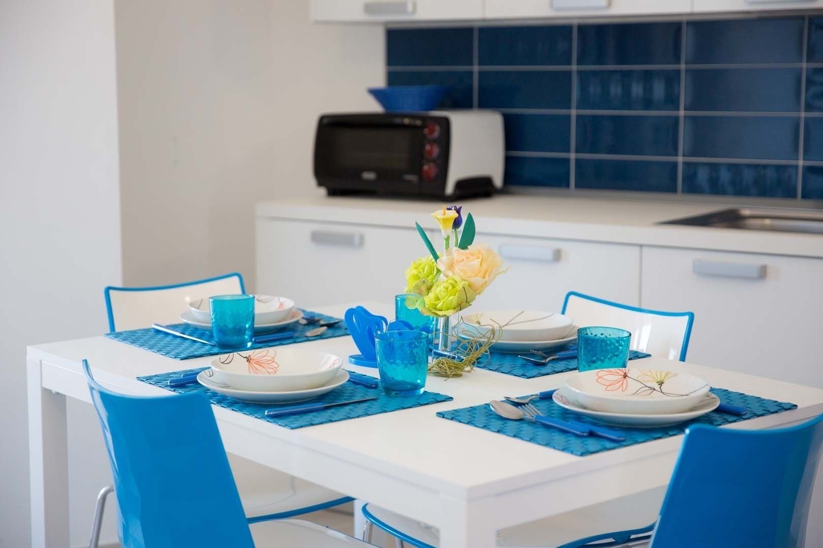 Ein Esstisch ist mit blauen Tischsets, Tellern und Gläsern gedeckt, in der Mitte steht eine Blumenvase.