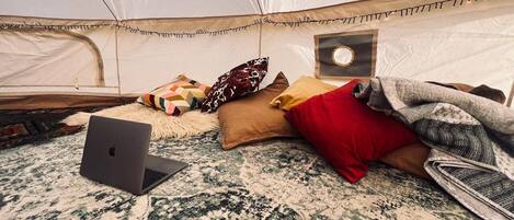 A tent interior with a laptop on a patterned rug, pillows, and a blanket.