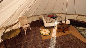 L’intérieur d’une tente, avec un lit, une chaise en osier, une petite table et un objet décoratif posé sur le sol.