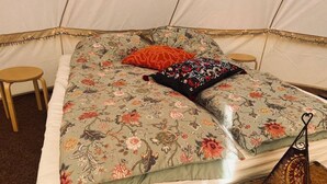 Un hébergement sous tente comprenant un lit avec une literie à motifs floraux, deux oreillers, une petite table ronde et un sac suspendu.