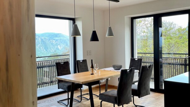 Un comedor moderno con mesa de madera y sillas negras, ventanas amplias con vistas a montañas y lámparas colgantes.