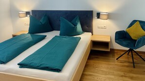 Una cama doble con ropa de cama color turquesa, dos lámparas fijadas a la pared, un sillón tapizado en azul oscuro con un cojín mostaza y una mesita de noche de madera.