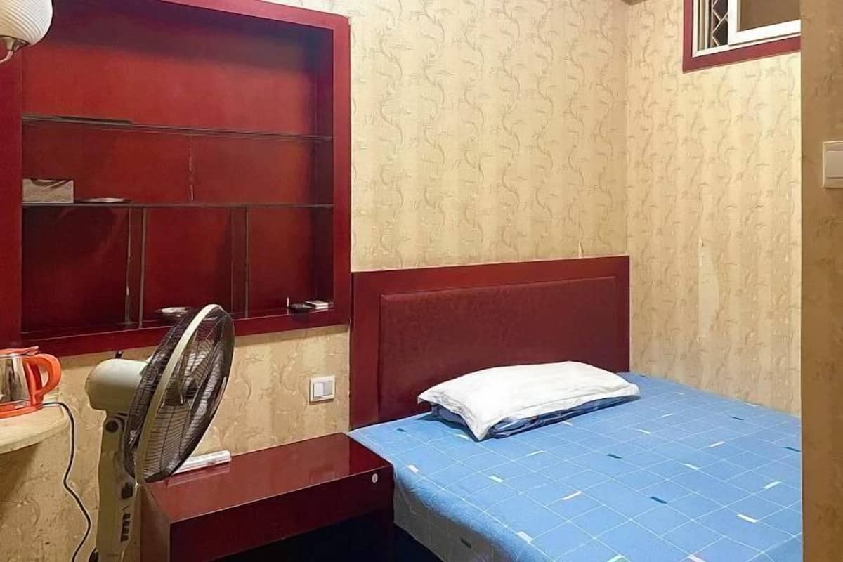 A small, neatly arranged room with a single bed, a nightstand, a red-framed window, and a wall-mounted fan.