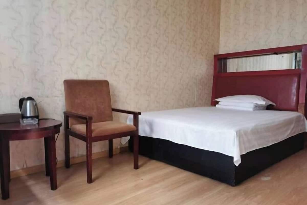 A hotel room with a bed, a wooden chair, a small table with a kettle, and patterned wallpaper.