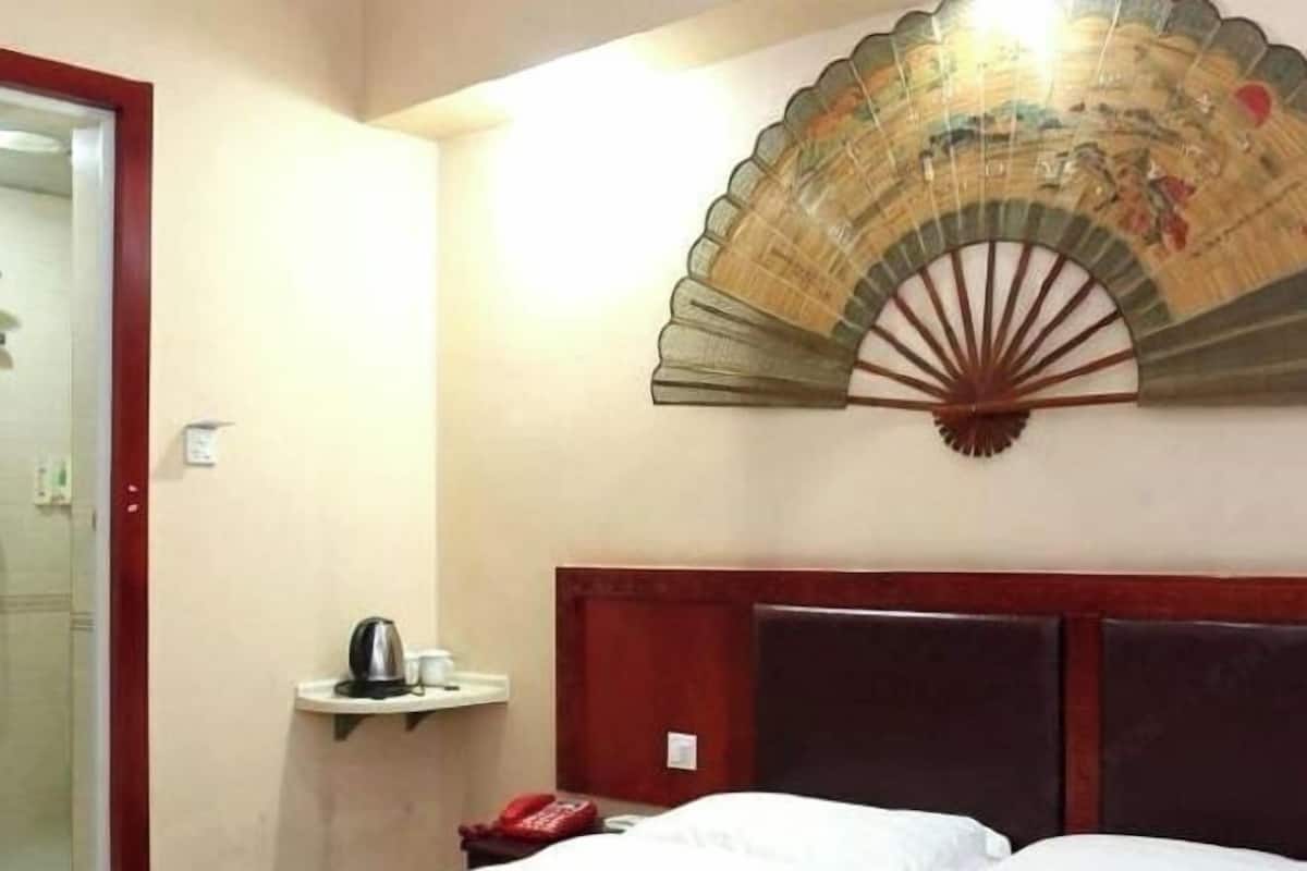 A hotel room with a bed, a bedside table with a kettle and a red telephone, and a decorative fan on the wall.