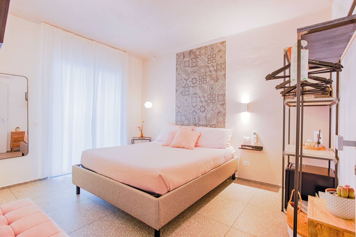 A bedroom with a bed, a bedside table, a hanging wardrobe, a wall with a geometric pattern, and a window with curtains.