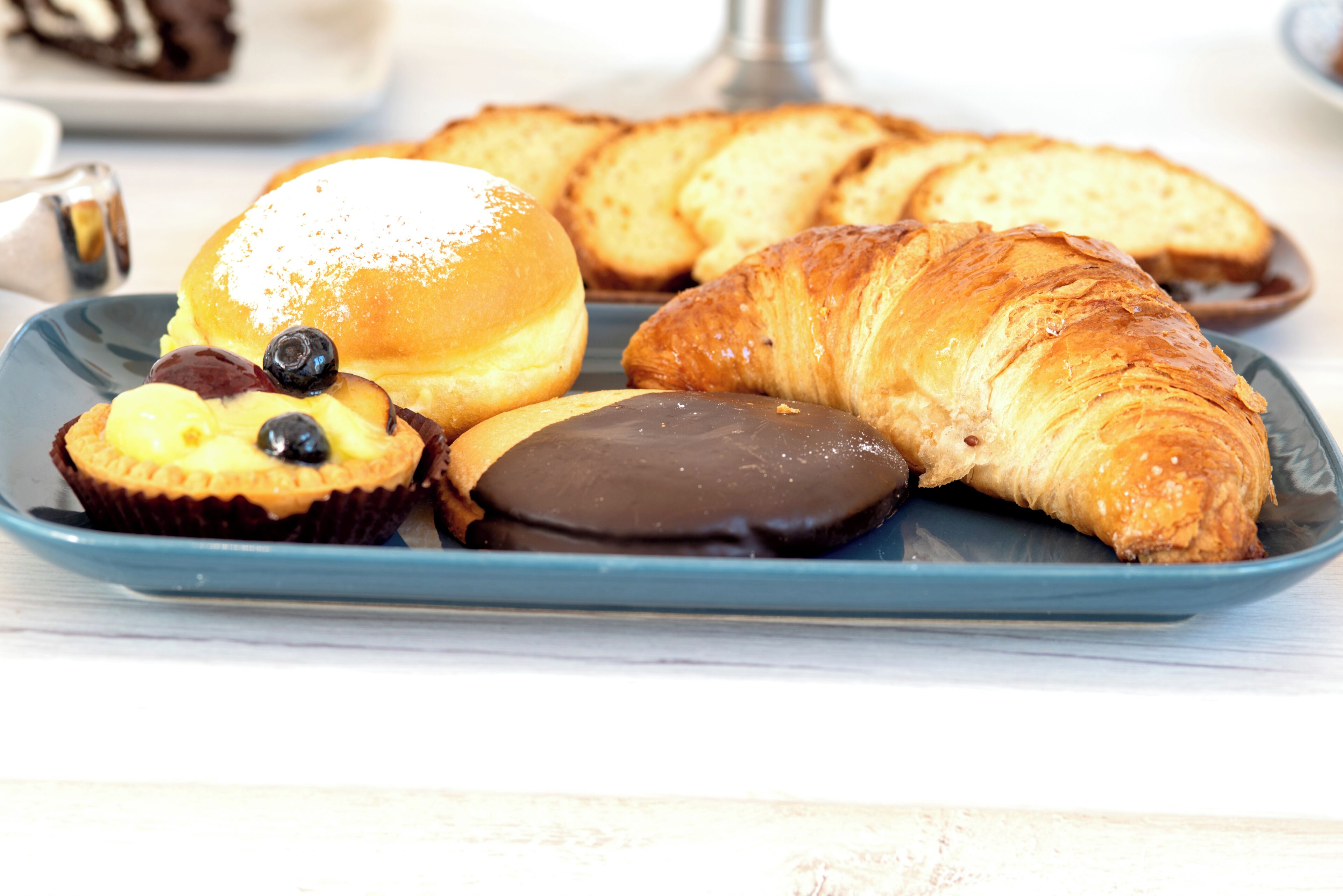 Un plato de pasteles que incluye un croissant, un dulce cubierto de chocolate, un muffin con arándanos y un donut con azúcar glas.