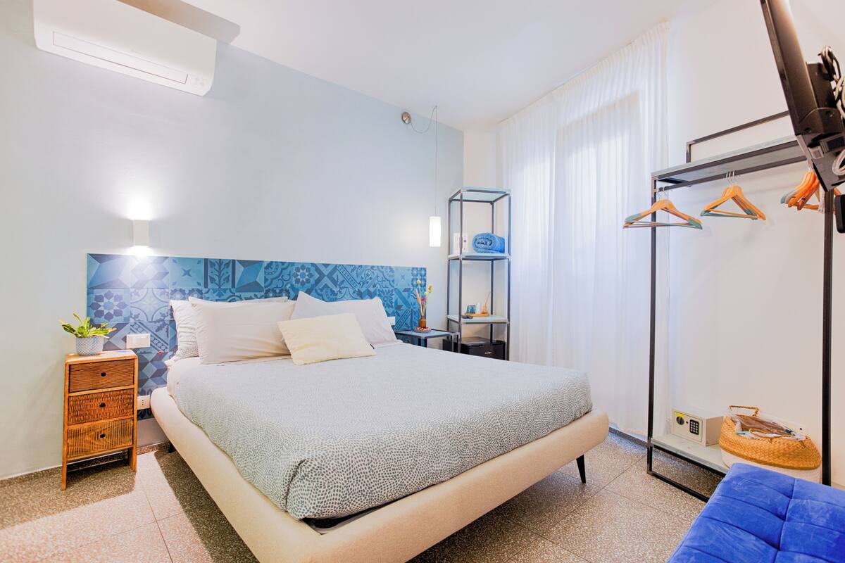 A bedroom with a bed, bedside table, lamp, and a wardrobe with hanging items.