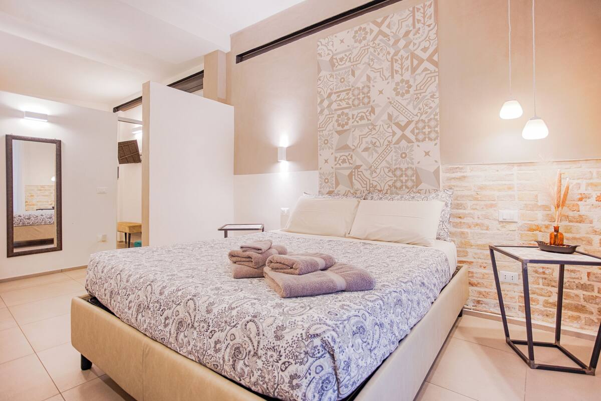 A modern bedroom with a large bed, a mirror, a wall-mounted light, and a small table with a vase.