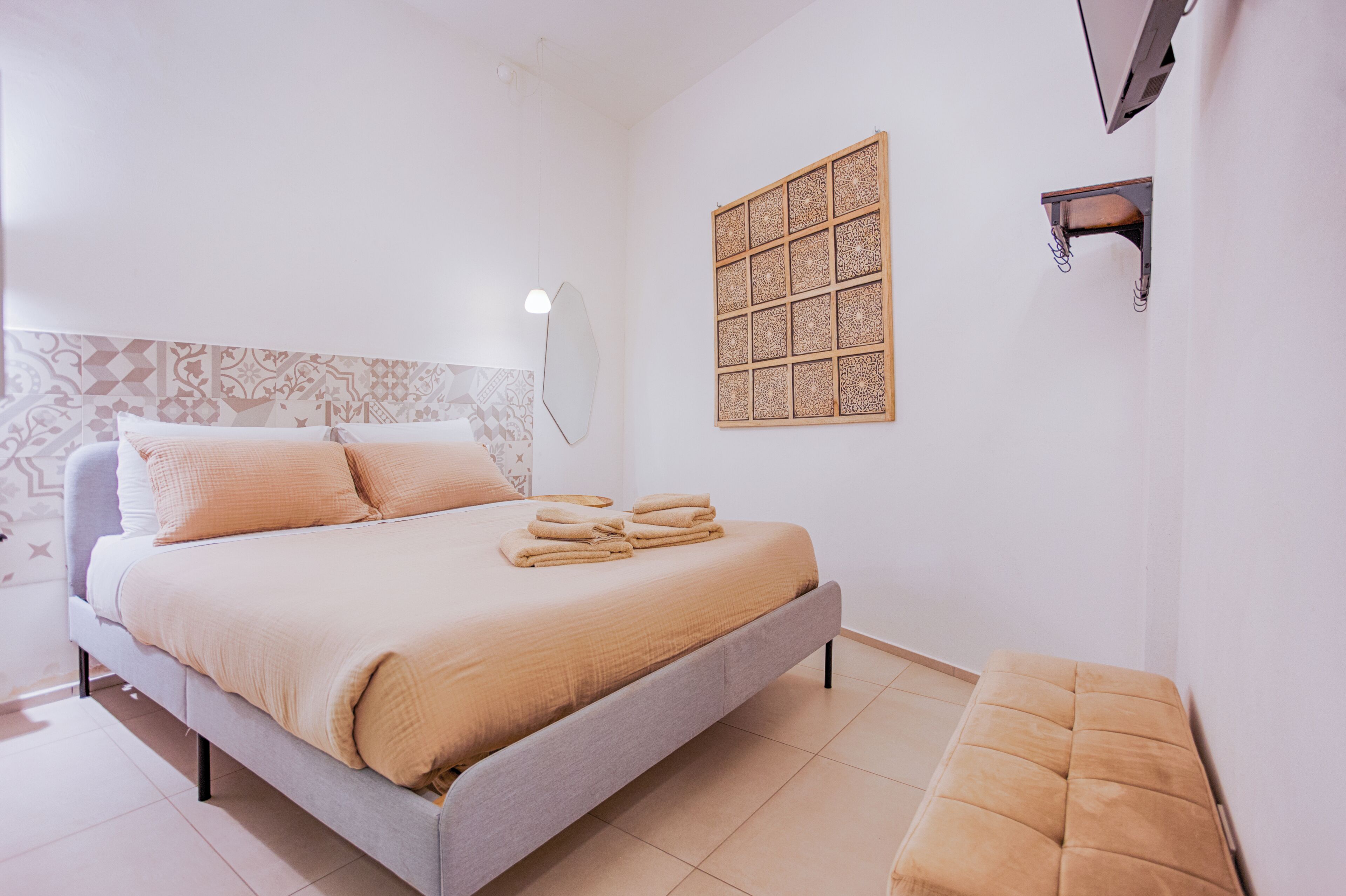 A neatly made bed with a headboard, a bedside table, a wall-mounted shelf, and a patterned wall decoration.