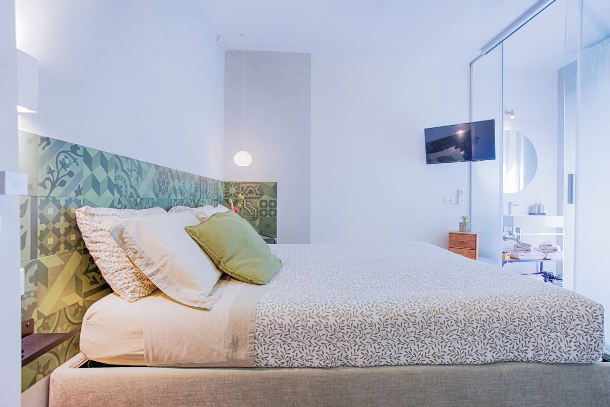 A neatly made bed with pillows, a bedside table with a lamp, a wall-mounted TV, and a bathroom with towels and toiletries.
