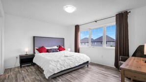 Quarto com cama, mesa de cabeceira, escrivaninha e janela com cortinas.