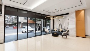 Un hall d’hôtel moderne avec une entrée en verre, un espace salon comprenant un canapé et des fauteuils, et un plafond doté d’un éclairage encastré.