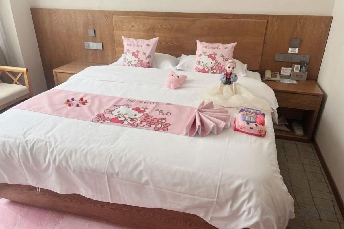 A neatly made bed with a pink and white bedspread, two pillows with a Hello Kitty design, and a Hello Kitty plush toy on the bed.