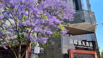 Un edificio de hotel con un letrero en un idioma extranjero, un árbol con flores moradas en primer plano, y un cielo despejado.