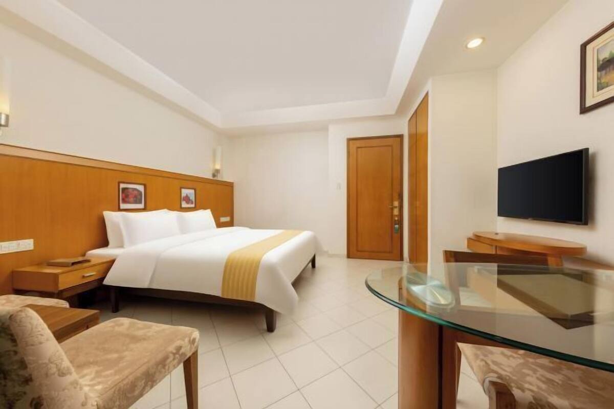 A hotel room with a bed, a TV, a glass table, and a wooden wardrobe.